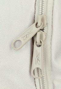 Dos tiradores de cremallera beige sobre un fondo de tela texturizada, posicionados muy cerca en una cremallera cerrada.