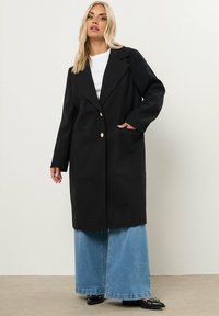 Abrigo largo negro con un amplio cuello, botones dorados de doble botonadura y dos bolsillos frontales, llevado sobre una blusa blanca y una falda de mezclilla.