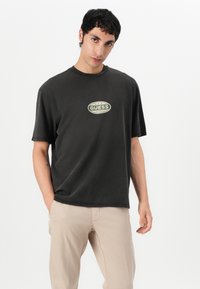 Svart t-skjorte i bomull med rund hals, kort ermer og et "Guess Originals"-logo i grønn og hvit på forsiden.