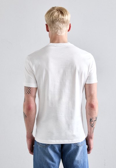 Tricou alb din bumbac cu guler clasic și mâneci scurte, asortat cu blugi albaștri. Tatuaje vizibile pe brațe.
