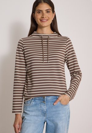 Femme souriante portant un haut à manches longues rayé marron et blanc avec des cordons et un jean bleu clair, une main dans la poche, sur fond neutre.