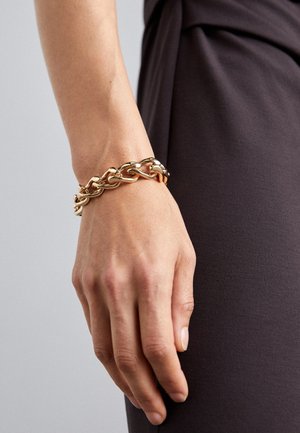Main portant un bracelet chaîne en or épais reposant à côté d'une manche sombre ou d'un vêtement sur un fond gris clair.