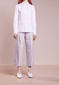Camicia bianca con colletto abbinata a pantaloni a vita alta di un tenue colore lavanda e tacchi bianchi a punta, in piedi su una superficie chiara.