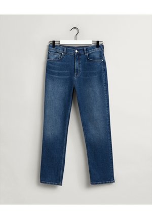 Jean en denim bleu avec une coupe droite, conçu avec cinq poches, quincaillerie argentée et décoloration subtile. Suspendu à un crochet noir sur un fond blanc.