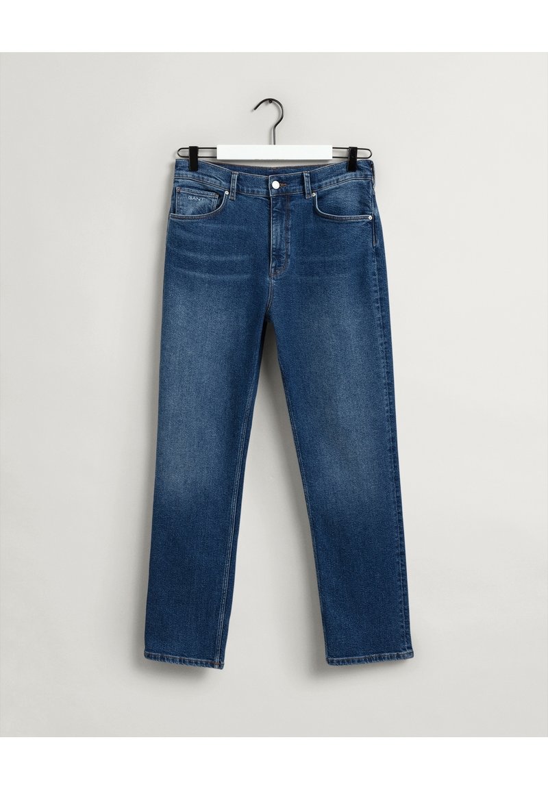 Modré džínové kalhoty se rovnými nohavicemi, designem pěti kapes, stříbrným kováním a mírným vyblednutím. Visí na černém háčku na bílém pozadí.