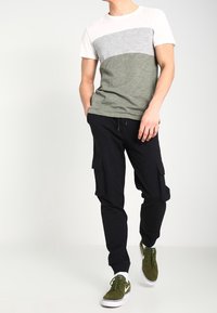 Camiseta de manga corta a rayas de color con un diseño en blanco, gris y verde oliva. Pantalones cargo negros con bolsillos laterales y zapatillas verdes.