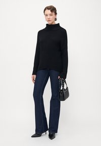 Kvinna klädd i en svart polotröja, mörkblå utsvängda jeans, svarta spetsiga klackar och håller en svart stunted handväska.