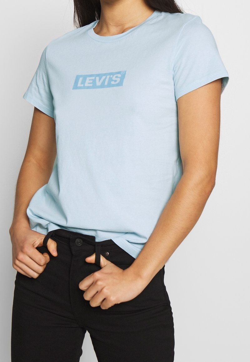 Personne portant un t-shirt Levi's bleu clair et un pantalon noir, les mains agrippant les passants de ceinture.