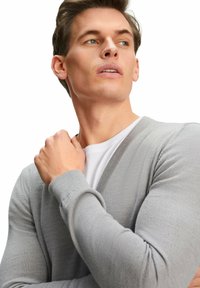 Giovane uomo con un cardigan grigio chiaro sopra una camicia bianca, che guarda di lato con la mano appoggiata vicino alla clavicola.