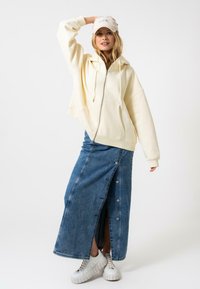 Sudadera cremosa con cremallera, falda de mezclilla larga con botones laterales y zapatillas blancas con suela texturizada, estilizadas con una gorra clara.