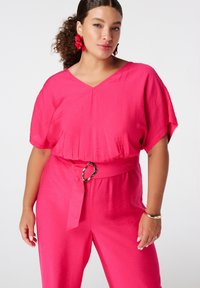 Combinaison rose vif avec une coupe décontractée, col en V, manches courtes, taille ceinturée avec un détail en anneau doré, et tissu texturé.