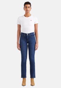 White short-sleeve t-shirt with a small red logo, paired with dark blue fitted jeans and beige ankle boots, standing against a neutral background.