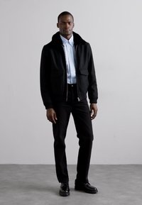 Svart zip-up jacka med en mjuk krage, kombinerad med en ljusblå skjorta och svarta byxor. Modellen står mot en neutral bakgrund.