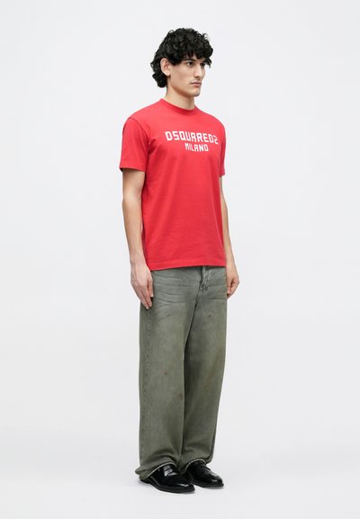 Punane lühikeste varrukatega T-särk, millel on "DSQUARED2 MILANO" logo, koos lahtise lõikega oliivroheliste teksade ja mustade jalanõudega.
