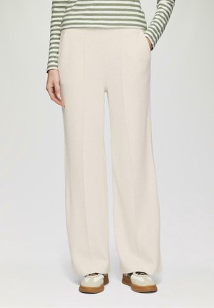 Pantalones anchos de color beige claro con bolsillos laterales y una textura suave, combinados con zapatillas blancas y marrones. Se ve una blusa a rayas verde y blanca.