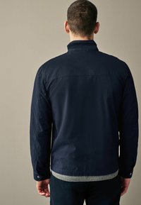 Granatowa kurtka z kołnierzem, zapinana na guziki przy mankietach, z prążkowanym szarym dołem, wykonana z gładkiej tkaniny, pokazana od tyłu.