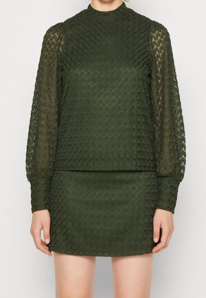 Vrouw die een donker groene kanten top met lange mouwen draagt en bijpassende korte rok met een zeshoekig patroon en pofmouwen.