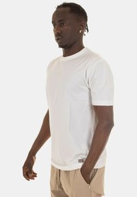 T-shirt bianco a maniche corte in cotone con colletto rotondo, dotato di una tasca sul petto discreta e un'etichetta decorativa sull'orlo.