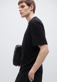 Μαύρο βαμβακερό μπλουζάκι με κοντά μανίκια, στρογγυλή λαιμόκοψη και άνετη εφαρμογή. Διαθέτει πλαϊνή τσέπη. Το μοντέλο κρατά μια μικρή μαύρη τσάντα.