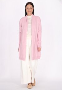 Ljusrosa cardigan med intrikat mönster, öppen front, ribbad mudd, bärs över en krämfärgad topp och vita vida byxor. Avslappnat fotbeklädnad i beige.