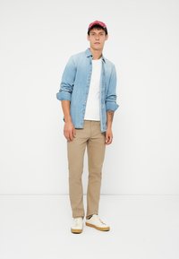 Ung mann står med beige bukser, hvite sneakers, hvit T-skjorte, ubottonert lyseblå denimskjorte og rød caps mot hvit bakgrunn.