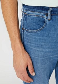 Blue denim jeans with a close-fitting design, featuring a classic five-pocket layout, light distressing, and contrasting orange stitching.