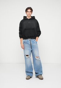 Musthavega kapuutsweater, millel on eesmine tasku ja katkised detailid, koos helesiniste laia säärega teksadega, millel on rebenenud alad ja lukuga detail.