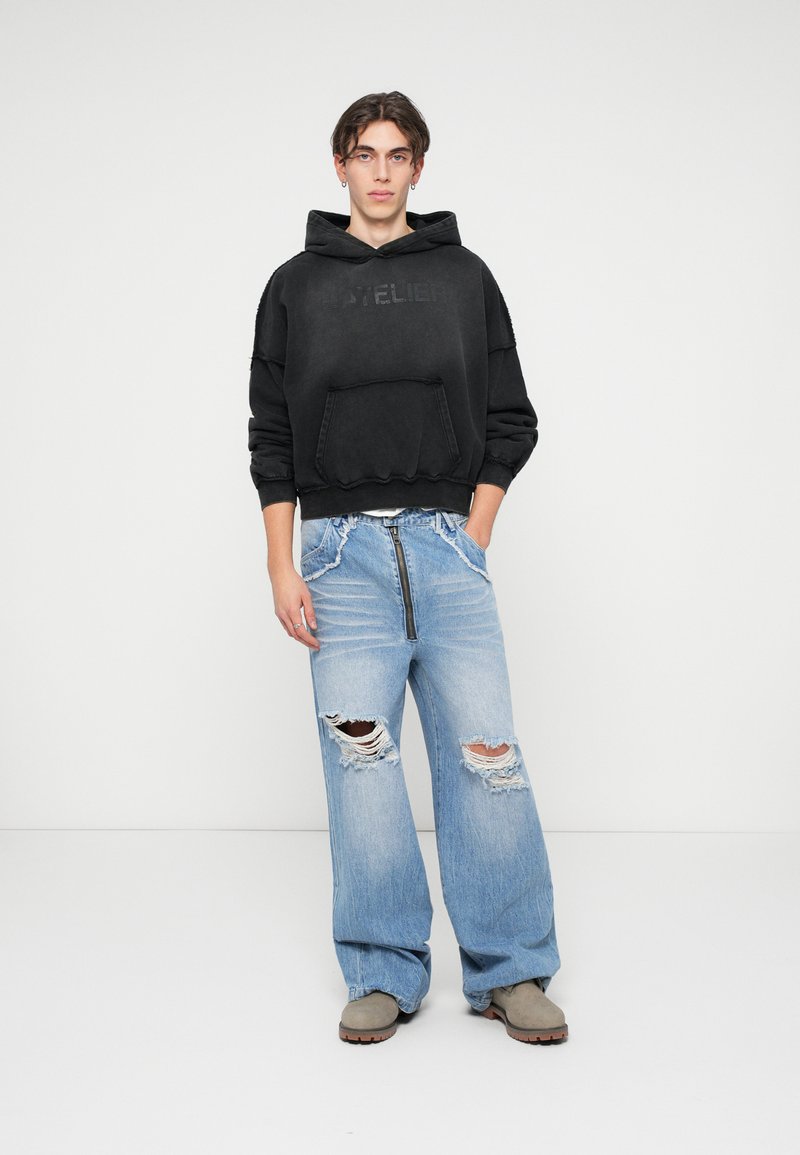 Sweatshirt preto com capuz, bolso frontal e detalhes desgastados, combinado com jeans de perna larga azul clara com áreas rasgadas e um zíper.