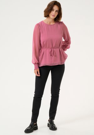 Rosa durchsichtige Bluse mit Kordelzug in der Taille, langen Puffärmeln mit gesmokten Bündchen, kombiniert mit figurbetonten schwarzen Hosen und klobigen Loafern.