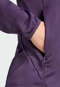 Tessuto viola texturizzato con polsini arricciati, dotato di una tasca laterale con chiusura a zip per una conservazione sicura.