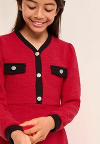 Rotes, strukturiertes Kleid mit schwarzem Besatz, langen Ärmeln, zwei Brusttaschen mit Knopfakzenten und dekorativen Steinen auf der Vorderseite.