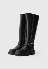 Par de botas de cuero negro hasta la rodilla con suelas tipo lug, cremallera lateral, paneles elásticos y correas decorativas en el tobillo con tachuelas y anillos en tono dorado.