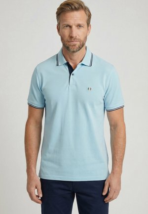 Man draagt een lichtblauw polo met korte mouwen met donkere afwerking en een klein geborduurd logo op de borst, gecombineerd met een donkere broek.