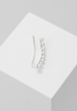 Boucle d'oreille argentée ornée de huit perles blanches rondes disposées en ligne courbe, posée sur une surface cubique blanche.