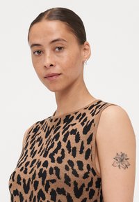 Αμάνικη μπλούζα σε καφέ πλέξη με μαύρο λεοπάρ σχέδιο, στρογγυλή λαιμόκοψη και λεία υφή; διαθέτει ένα λουλουδάτο τατουάζ στο χέρι.