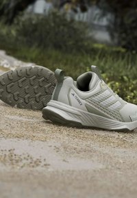 Ανοιχτό γκρι παπούτσια πεζοπορίας Adidas Terrex σε χερσαίο μονοπάτι με ανθεκτική σόλα, θολό πράσινο φόντο εξωτερικού χώρου.