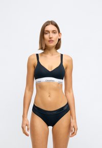 Černý bavlněný bralet s V-výstřihem a nastavitelnými ramínky, s bílým logem Calvin Klein na pásu a odpovídajícími černými kalhotkami.