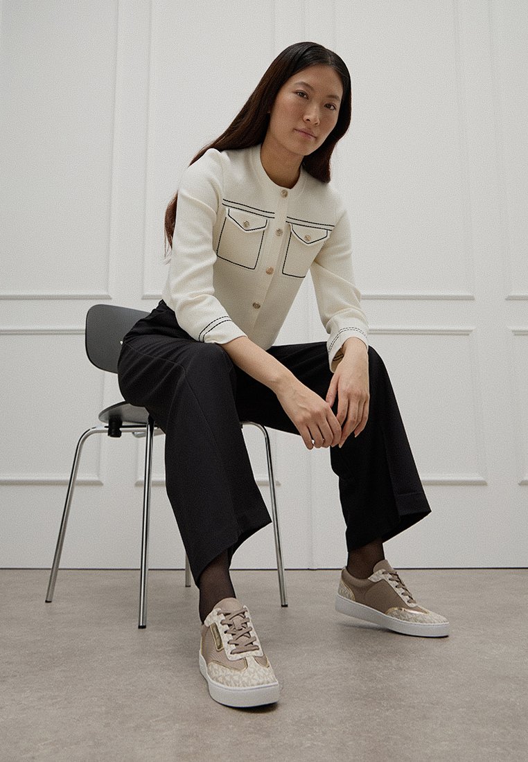 Cardigan crème avec deux poches poitrine et contour noir, associé à un pantalon noir ample et des baskets beiges avec des accents texturés.