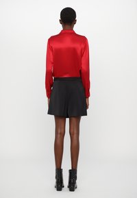 Rotes Satin-Hemd mit langen Ärmeln, schwarze gefältete Shorts und schwarze Knöchelstiefel mit silbernen Beschlägen. Glatte Textur, taillierte Silhouette, modernes Design.