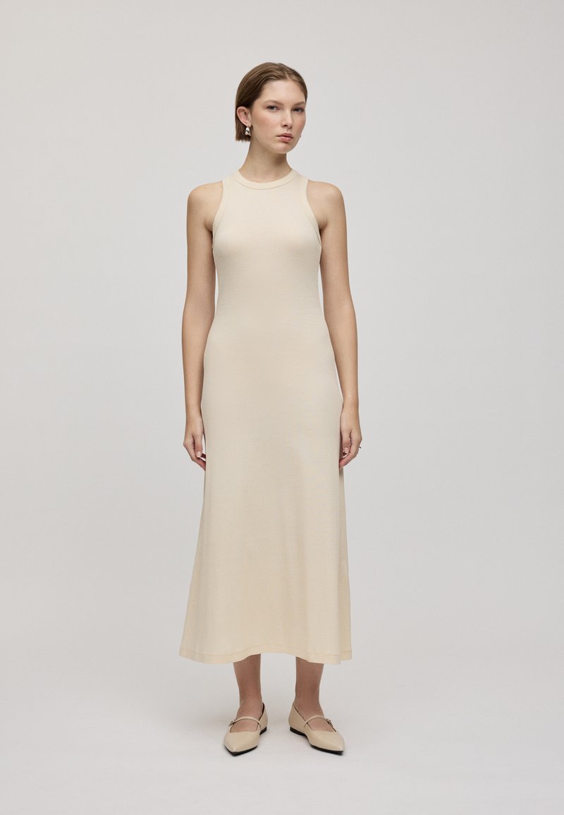 Abito senza maniche di colore crema, aderente, con lunghezza midi e texture liscia. Modella abbinato a scarpe slip-on beige.