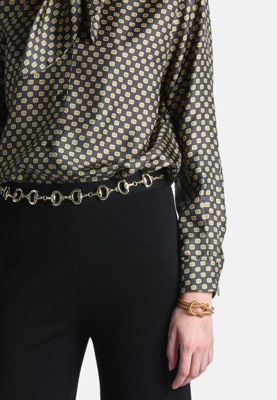 Femme portant un chemisier noir à manches longues à motifs, un pantalon noir, une ceinture chaîne en or et un bracelet torsadé en or à son poignet droit.