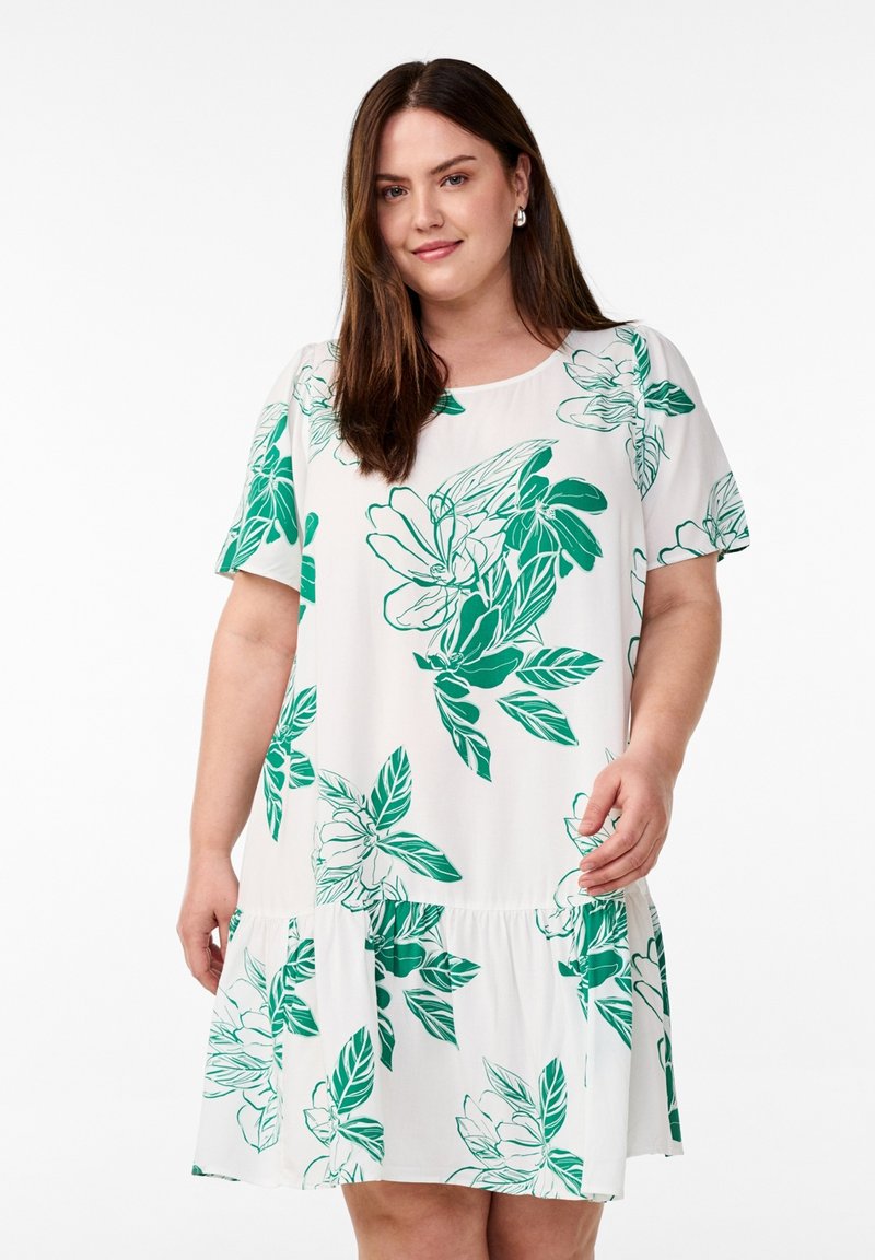 Vrouw met bruin haar die een witte jurk met korte mouwen en groen bloemenpatroon draagt, staande tegen een witte achtergrond.