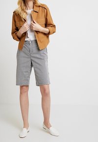 Giacca marrone con bottoni a pressione, t-shirt bianca, pantaloni corti a righe grigio e bianchi, e scarpe bianche senza lacci. Look casual, vestibilità rilassata; nessun motivo visibile.