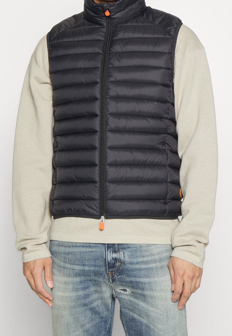 Chaleco acolchado negro con patrón de rombos, cuello alto y tirador de cremallera naranja. Lleva encima un sudaderón beige claro, combinado con jeans desgastados.