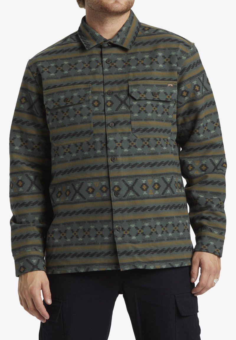 Knopshirt in het groen, met geometrische patronen in bruin en geel. Twee borstzakken, lange mouwen en een kraag.