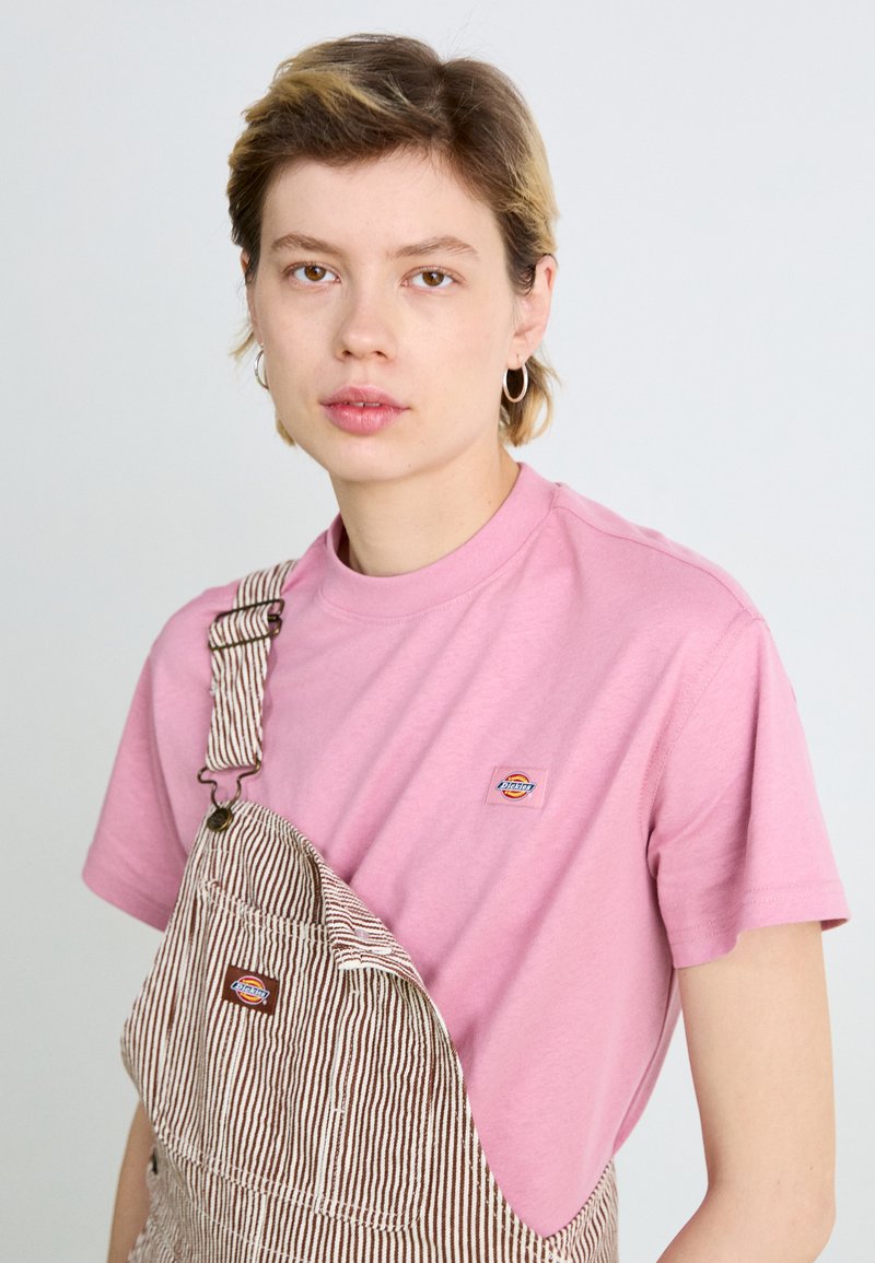 T-shirt di cotone rosa abbinata a una salopette a righe marroni e bianche. I dettagli notevoli includono un piccolo logo sul petto e bretelle regolabili.