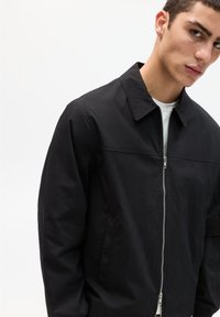 Schwarze leichte Bomberjacke mit Reißverschluss, Umlegekragen und seitlichen Taschen. Glatte Textur und minimale Designmerkmale.