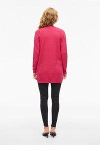 Long-sleeve cardigan in bright pink knit material, featuring a rounded hem and ribbed cuffs, paired with black skinny jeans and heels.