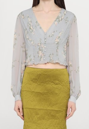 Blouse fleurie gris clair à manches longues et col en V, avec des plis et des boutons à l'avant, associée à une jupe vert olive froissée.