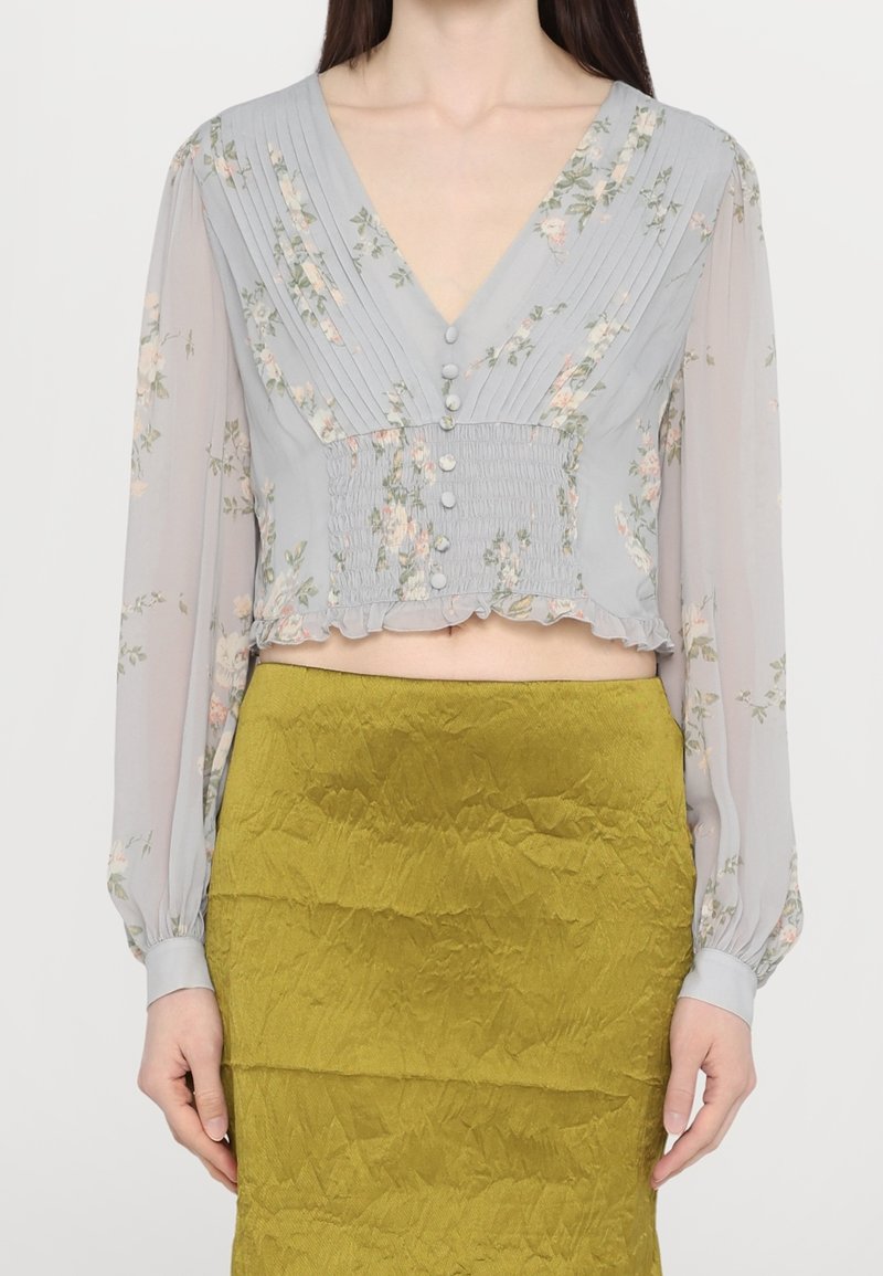 Blouse fleurie gris clair à manches longues et col en V, avec des plis et des boutons à l'avant, associée à une jupe vert olive froissée.